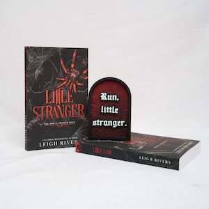 May include: Two books titled Little Stranger and Little Liar by Leigh Rivers, featuring black covers with red spider and web graphics. Between them sits an arched, black-framed sign with a textured red background and the white text Run, little stranger.