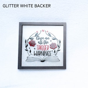 May include: A framed art print with a white glitter background. The print features a black and white illustration of an open book with roses and vines. The text "Give me all the trigger warnings" is written in red.
