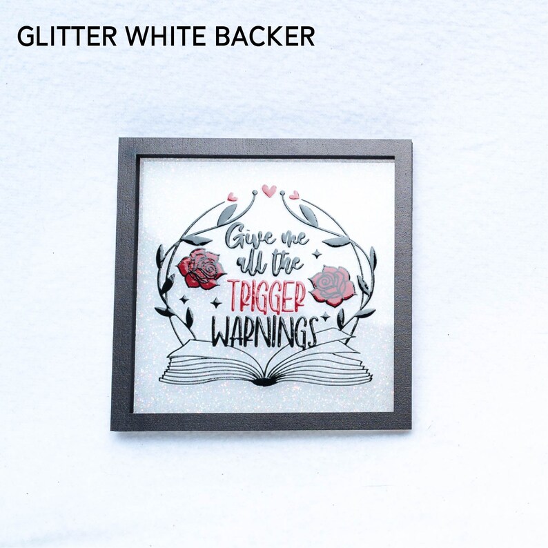May include: A framed art print with a white glitter background. The print features a black and white illustration of an open book with roses and vines. The text "Give me all the trigger warnings" is written in red.