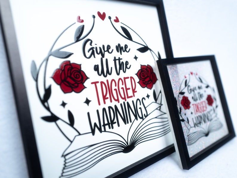 May include: A framed print with a white background and black text that reads "Give me all the trigger warnings". The text is surrounded by a black and white illustration of a book, roses, and vines. Two red hearts are at the top of the image.
