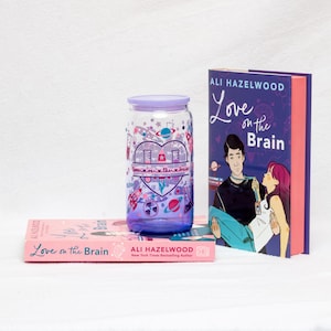 May include: A purple-tinted glass tumbler with a lid, decorated with a heart-shaped design and the words "love on the brain." Two books with the title "Love on the Brain" are also visible, one pink and one purple.