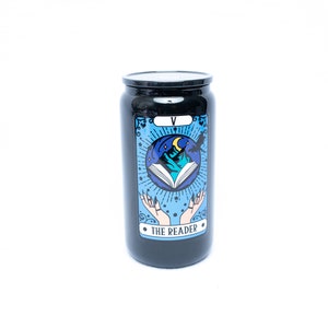 May include: Black candle jar with a tarot card design. The card depicts a blue and white illustration of a book, mountains, and a crescent moon. The text on the card reads "The Reader" and "V".