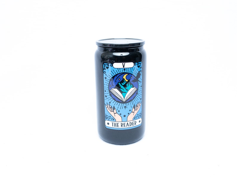 May include: Black candle jar with a tarot card design. The card depicts a blue and white illustration of a book, mountains, and a crescent moon. The text on the card reads "The Reader" and "V".