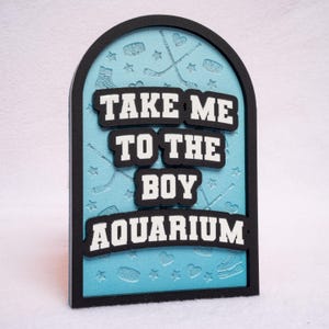May include: A decorative sign with a black frame and a light blue background. The sign reads "TAKE ME TO THE BOY AQUARIUM" in white, raised lettering. Hockey-themed icons are embossed on the background.