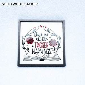 May include: A framed print with a white background and black and silver lettering. The print features a book with roses and vines around it. The text reads "Give me all the trigger warnings".