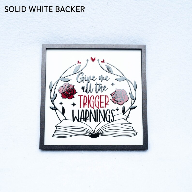 May include: A framed print with a white background and black and silver lettering. The print features a book with roses and vines around it. The text reads "Give me all the trigger warnings".