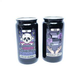 May include: Two black canisters with tarot card designs. The left canister has a skull and crossbones with the text "Dubious Consent", "Torture", "Knife Play", "Stalking", and "Trigger Warnings". The right canister has two hands with the text "Morally Grey".