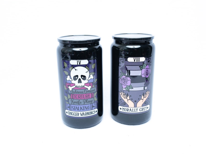 May include: Two black canisters with tarot card designs. The left canister has a skull and crossbones with the text "Dubious Consent", "Torture", "Knife Play", "Stalking", and "Trigger Warnings". The right canister has two hands with the text "Morally Grey".