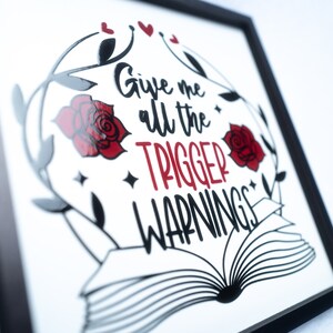 May include: A framed print with a black frame and a white background. The print features a black and white illustration of an open book with a red rose and leaves on either side. The text "Give me all the trigger warnings" is written in red and black.