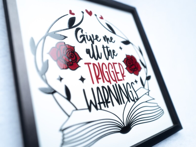 May include: A framed print with a black frame and a white background. The print features a black and white illustration of an open book with a red rose and leaves on either side. The text "Give me all the trigger warnings" is written in red and black.