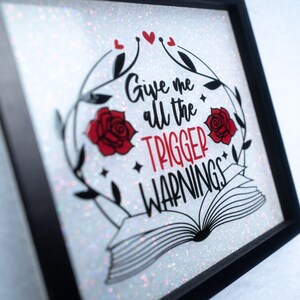 May include: A white glitter framed sign with black lettering that reads "Give me all the trigger warnings". The sign features two red roses and a black and white illustration of an open book.