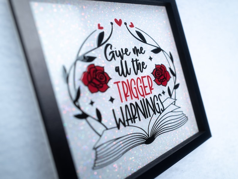 May include: A white glitter framed sign with black lettering that reads "Give me all the trigger warnings". The sign features two red roses and a black and white illustration of an open book.