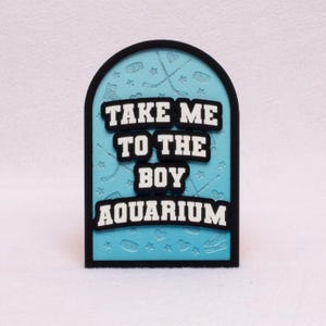 May include: A decorative sign with a black border and a light blue background. The sign features the text "TAKE ME TO THE BOY AQUARIUM" in white letters with black outlines. Hockey-themed graphics are embossed on the background.