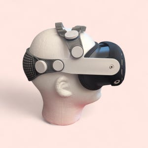 May include: A virtual reality headset with a white and gray frame, black eye covers, and adjustable straps. The headset is mounted on a white foam head, showcasing its design and fit. The design includes circular adjustment knobs and a textured strap.