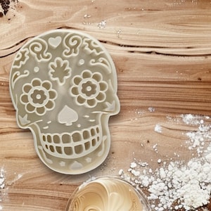 May include: White plastic sugar skull cookie cutter with intricate floral and swirl designs.