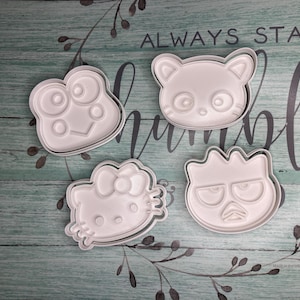 Może przedstawiać: Cztery białe plastikowe foremki do ciasteczek w kształcie żaby, kota, postaci Hello Kitty i potwora z marszczonym czołem. Formy znajdują się na zielono-białej drewnianej powierzchni.