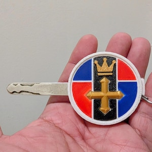 May include: A custom key with a decorative emblem. The emblem is round, featuring a gold cross and crown on a background of red, black, and blue segments. The key is held by a hand.