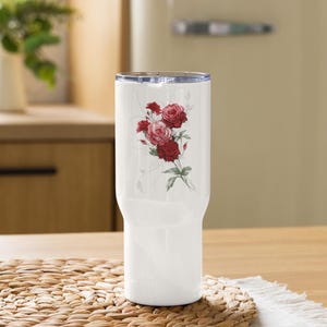 May include: White insulated tumbler with a floral design featuring red and pink roses. The tumbler has a clear lid and is sitting on a woven mat. The floral design is centered on the tumbler.