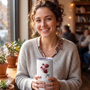 May include: A white insulated tumbler featuring a floral design with red roses and green leaves. The tumbler is held by a person wearing a gray sweater and a floral print shirt. The background includes potted plants and a window.