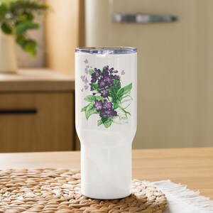 May include: A white insulated tumbler featuring a floral design of purple violets and green leaves. The tumbler has a clear lid and is designed to keep beverages hot or cold. The floral design is a watercolor style.