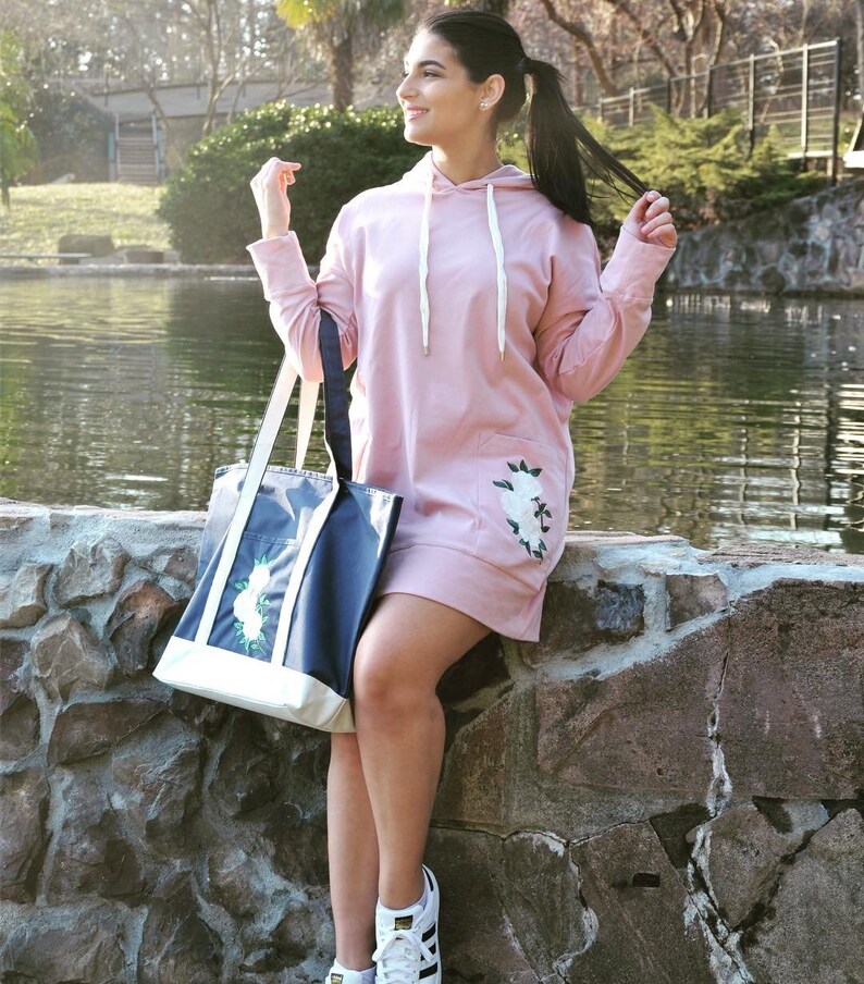 May include: A pink hooded sweatshirt with a floral embroidered pocket and a white drawstring. The sweatshirt is worn by a person holding a navy blue tote bag with a white floral design.