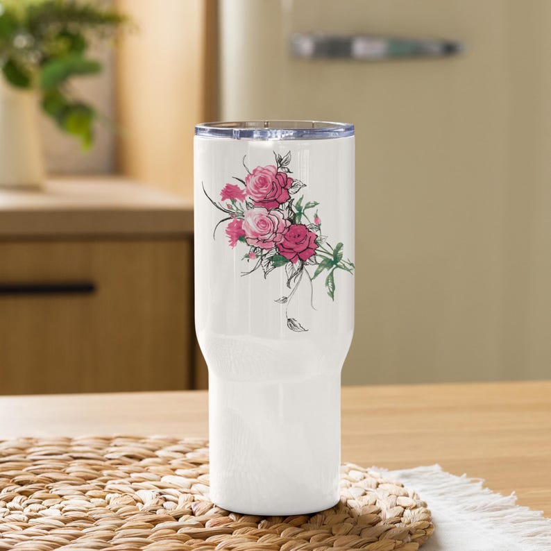 May include: White insulated tumbler featuring a floral design of pink and red roses with green leaves. The tumbler has a clear lid and is designed to keep beverages cold or hot. The tumbler is sitting on a woven mat.