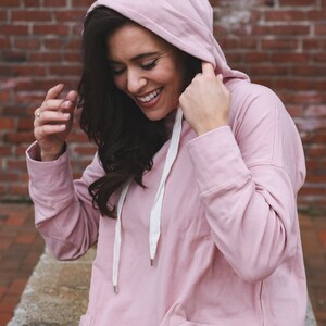 May include: A light pink hooded sweatshirt with a front pocket and white drawstring. The sweatshirt features a floral embroidery on the front pocket. The model is wearing the sweatshirt and smiling.