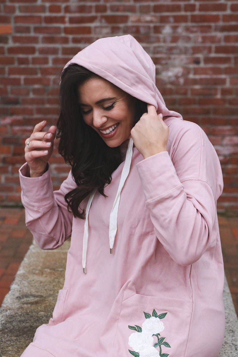 May include: A light pink hooded sweatshirt with a front pocket and white drawstring. The sweatshirt features a floral embroidery on the front pocket. The model is wearing the sweatshirt and smiling.