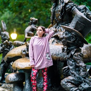 May include: A person wearing a light purple hoodie with a floral pocket and red floral leggings stands near the Alice in Wonderland statue. The statue features Alice, the Mad Hatter, and other characters.