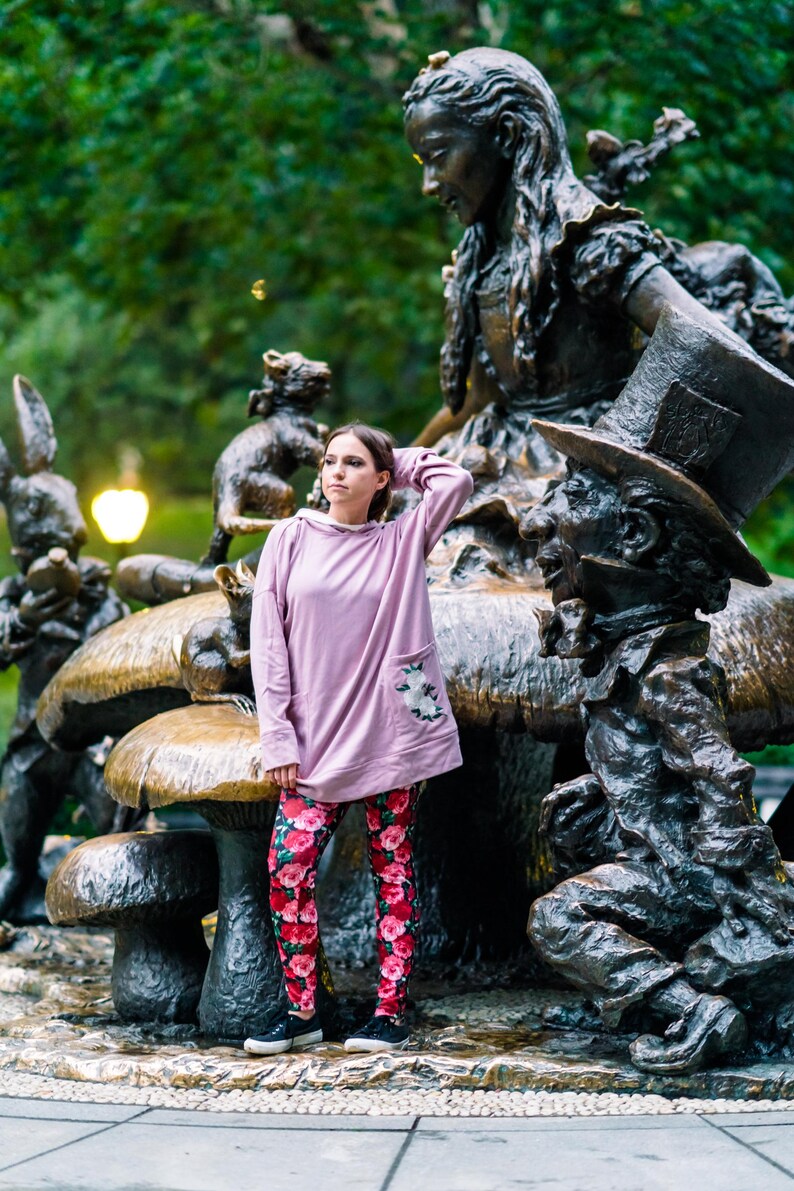 May include: A person wearing a light purple hoodie with a floral pocket and red floral leggings stands near the Alice in Wonderland statue. The statue features Alice, the Mad Hatter, and other characters.