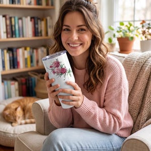 May include: White tumbler with a floral design featuring pink and red roses. The tumbler is held by a person wearing a pink sweater and blue jeans. The background includes a bookshelf, a window, and a cat.