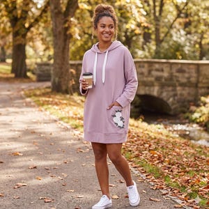 May include: A mauve hooded sweatshirt dress with a white drawstring and a floral embroidered pocket. The dress is knee-length and has long sleeves. The person is wearing white sneakers and holding a coffee cup.