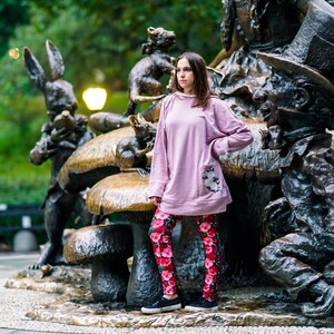 May include: A person wearing a light pink sweatshirt with a floral pocket and red floral leggings stands in front of an Alice in Wonderland bronze statue. The statue features the Mad Hatter, the White Rabbit, and other characters.