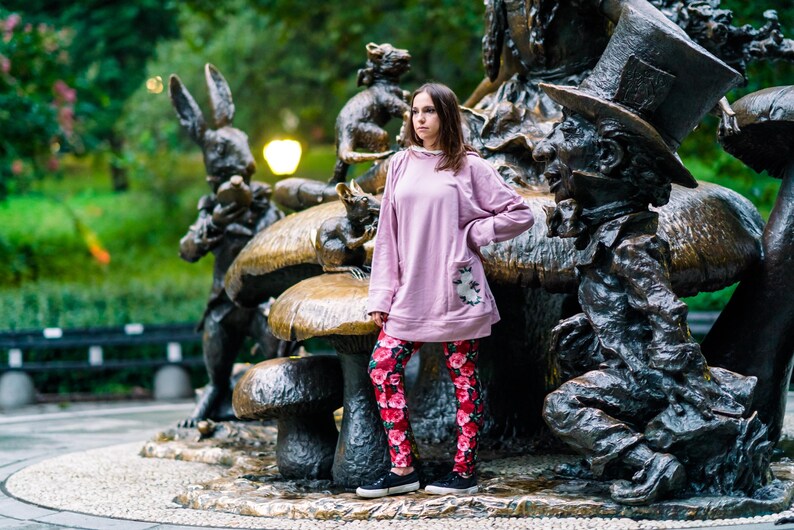 May include: A person wearing a light pink sweatshirt with a floral pocket and red floral leggings stands in front of an Alice in Wonderland bronze statue. The statue features the Mad Hatter, the White Rabbit, and other characters.