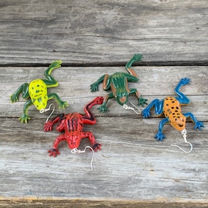 Pode incluir: Quatro brincos em forma de rã em várias cores: amarelo com manchas pretas, verde e castanho, vermelho com riscas pretas e laranja com membros azuis e manchas pretas. Cada brinco tem um gancho prateado.