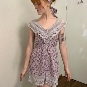 May include: A purple and white floral print mini dress with a white lace trim and a button-up front. The dress has short sleeves and a fitted bodice.