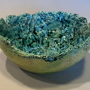 Puede incluir: Un cuenco de cerámica verde con un borde texturizado irregular y un esmalte moteado. El cuenco tiene una forma orgánica única y una apariencia rústica.