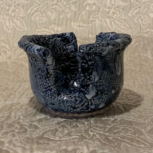 May include: A small, blue ceramic bowl with a textured, crackled glaze. The bowl has a unique, organic shape with a wide, open top and a rounded bottom.