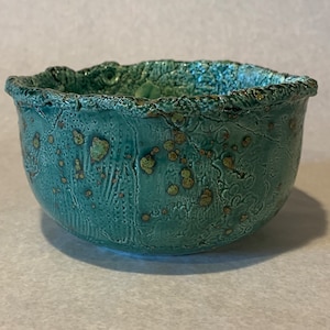 Pode incluir: Uma tigela de cerâmica azul-esverdeada com uma superfície texturizada e manchas douradas. A tigela tem uma borda ligeiramente irregular.