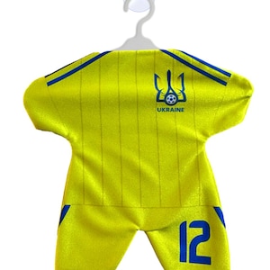 Peut inclure: Un maillot de football jaune avec des détails bleus, dont le numéro 12 sur le short. Le maillot arbore les armoiries ukrainiennes et le mot "UKRAINE". L'ensemble est suspendu à une ventouse blanche.