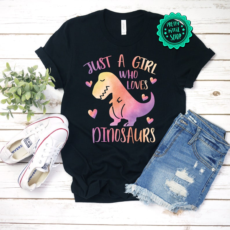 以下が含まれることがあります： 水彩の恐竜のグラフィックと「Just a girl who loves dinosaurs」というテキストが書かれた黒いTシャツです。