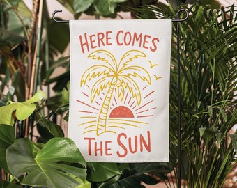 May include: A white rectangular garden flag featuring a graphic illustration of a yellow palm tree and a red sun. The text "HERE COMES THE SUN" is printed in red capital letters above and below the illustration, with two small yellow birds.