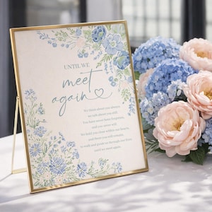 May include: A gold-framed sign with the words "UNTIL WE meet again" and floral decorations in blue. A bouquet of blue hydrangeas and pink peonies is next to the sign. The sign's text includes a heartfelt message.