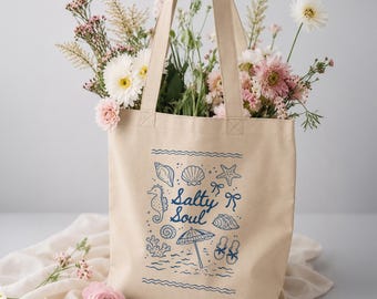 Pode incluir: Uma sacola de lona bege com um desenho azul com tema de praia e as palavras "Salty Soul". A sacola é preenchida com flores rosa e brancas. O gráfico inclui conchas, um cavalo-marinho e um guarda-sol.