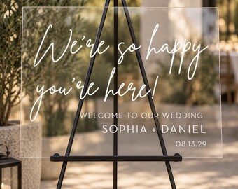 May include: A rectangular clear acrylic wedding sign displayed on a black metal tripod easel. The sign features white text that reads: We're so happy you're here! WELCOME TO OUR WEDDING SOPHIA + DANIEL 08.13.29. The sign is transparent and rigid.