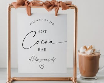 May include: A rectangular copper pipe frame holds a white sign with black text that reads WARM UP AT THE HOT Cocoa BAR Help yourself and a small heart icon. The sign is attached to the top bar with two brown fabric ribbons. A glass mug of cocoa.