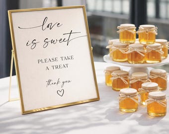 May include: A gold-framed sign with off-white paper featuring black text that reads love is sweet PLEASE TAKE A TREAT thank you and a heart symbol, displayed next to a white tiered stand holding multiple small glass jars filled with golden honey.