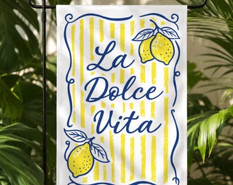 May include: A rectangular garden flag featuring a white background with vertical yellow stripes. The text La Dolce Vita is printed in blue script in the center. The design includes blue-outlined yellow lemons with leaves and a blue border.