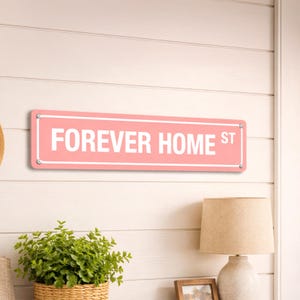 May include: A rectangular, pink metal sign with the words "FOREVER HOME ST" in white lettering. The sign is mounted on a white, horizontal-planked wall with silver-colored screws. A lamp and greenery are visible below.
