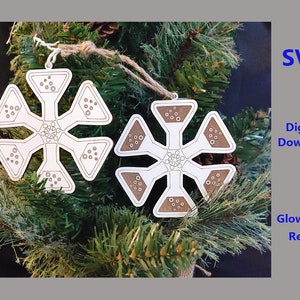 May include: Two white snowflake ornaments with a brown outline and a pattern of small circles. The ornaments are hanging on a green pine branch.